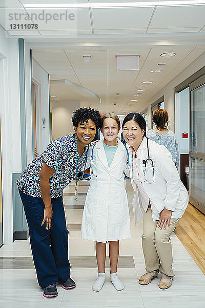 Ganzfigurenporträt von fröhlichen Ärztinnen mit Mädchen im Laborkittel im Krankenhauskorridor