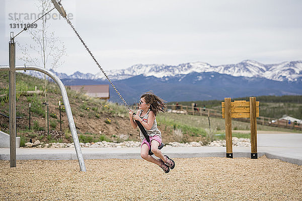 Glückliches Mädchen schwingt auf Kettenschaukel gegen Berge im Park