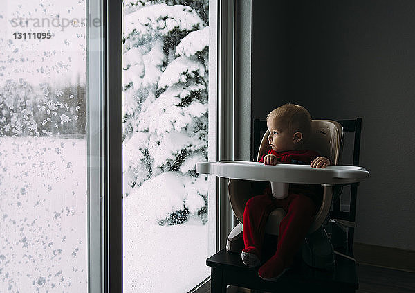 Kleiner Junge schaut durchs Fenster  während er im Winter zu Hause auf einem Hochstuhl sitzt