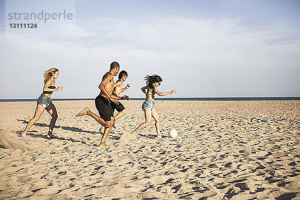 Freunde spielen Fussball auf Sand am Strand gegen den Himmel