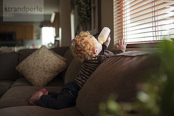 Seitenansicht eines kleinen Jungen  der Milch trinkt  während er sich zu Hause auf dem Sofa am Fenster entspannt