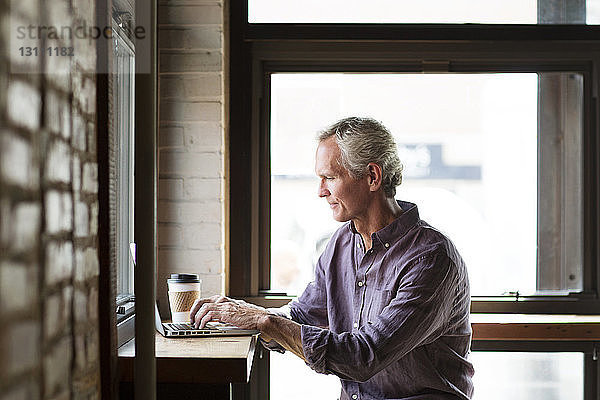 Reifer Mann mit Laptop am Tisch  der im Café an Glasfenstern sitzt