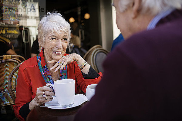 Glückliche ältere Frau hält Kaffeetasse in der Hand  während sie mit einem Mann im Straßencafé sitzt