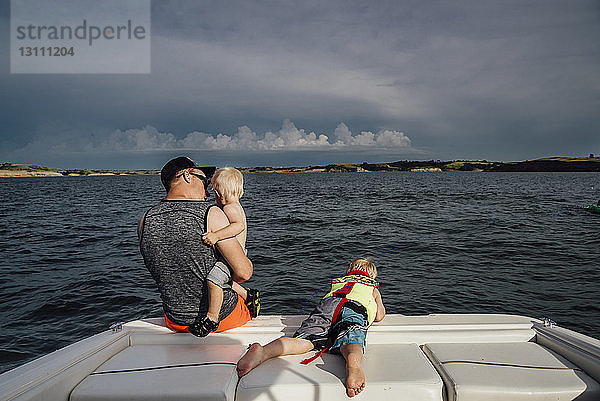 Rückansicht eines Vaters mit Söhnen  die im Boot auf dem Meer vor bewölktem Himmel reisen