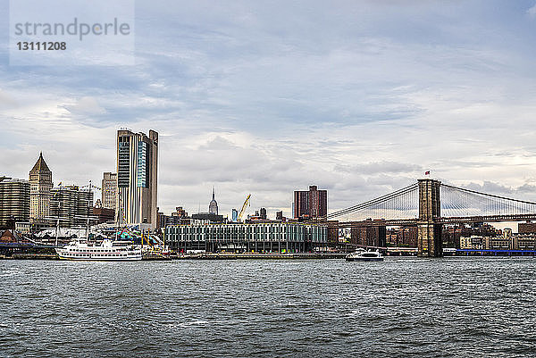 Brooklyn Bridge über den East River bei Stadt gegen bewölkten Himmel