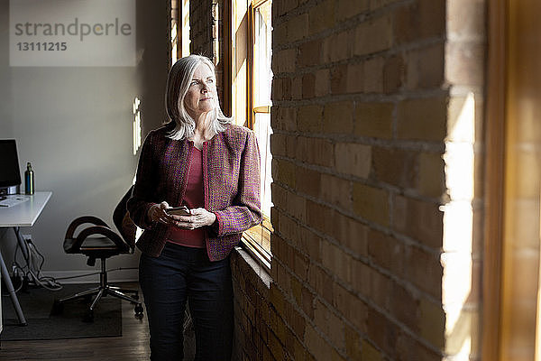 Nachdenkliche reife Geschäftsfrau schaut durchs Fenster  während sie im Kreativbüro ein Smartphone in der Hand hält