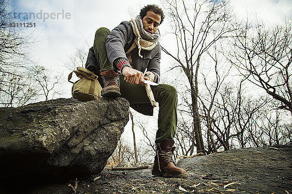 Niedrigwinkelansicht eines Mannes  der auf einem Felsen sitzt und Holz gegen den Himmel schneidet