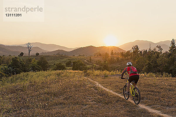 Rückansicht eines Mountainbike fahrenden Mannes auf dem Feld gegen den Himmel bei Sonnenuntergang