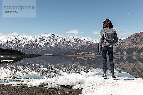 Frau in voller Länge auf Schnee am Fluss stehend  während sie gegen Berge und Himmel blickt