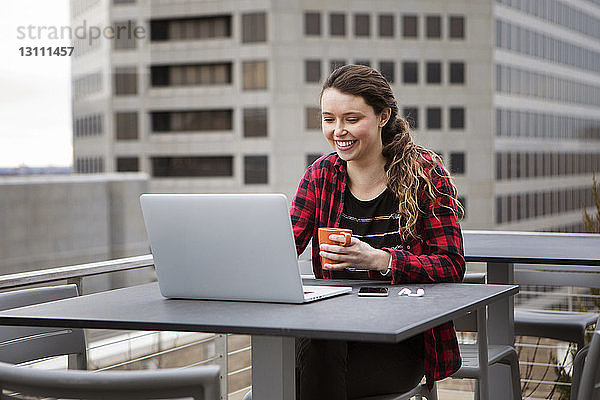 Lächelnde junge Frau benutzt Laptop-Computer  während sie auf der Terrasse eines Cafés sitzt