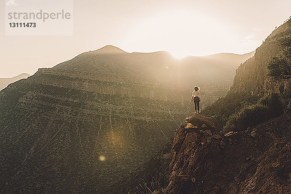 Rückansicht einer Frau  die den Berg betrachtet  während sie bei Sonnenuntergang auf einem Felsen gegen den Himmel steht