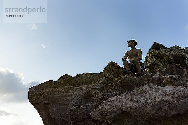 Tiefwinkelansicht eines Wanderers  der auf einer Felsformation vor blauem Himmel sitzt