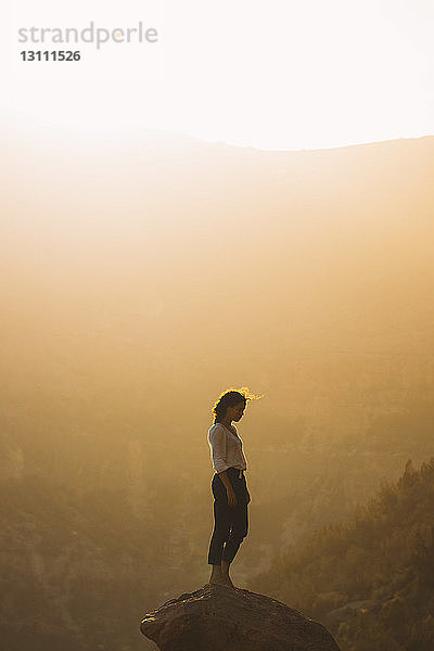 Seitenansicht einer Frau  die bei Sonnenuntergang auf einem Felsen gegen den Himmel steht