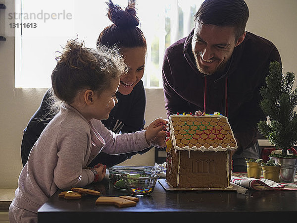 Glückliche Eltern sehen Tochter beim Dekorieren des Lebkuchenhauses auf dem Tisch