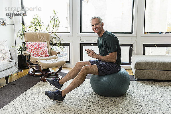 Porträt eines reifen Mannes in voller Länge  der zu Hause auf einem Fitnessball vor dem Fenster sitzt