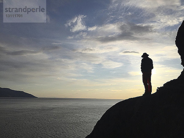 Silhouettenmann steht bei Sonnenuntergang auf felsigem Berg am Meer
