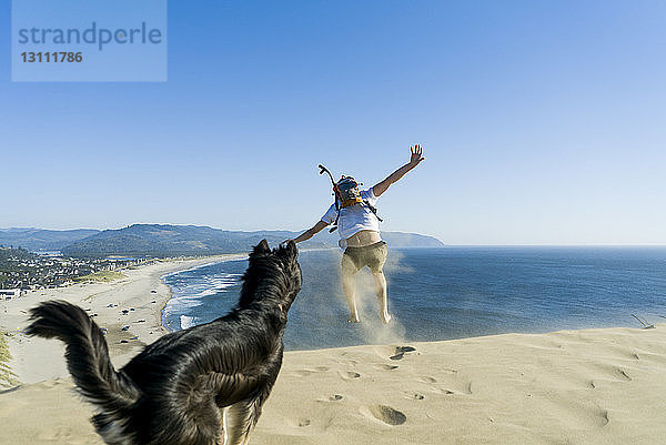 Verspielter Mann springt  während der Hund am Strand vor klarem blauen Himmel steht