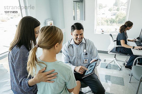 Hochwinkelansicht eines Kinderarztes  der Mutter und Tochter ein Röntgenbild auf einem Tablet-Computer zeigt  während die Ärztin im Hintergrund arbeitet