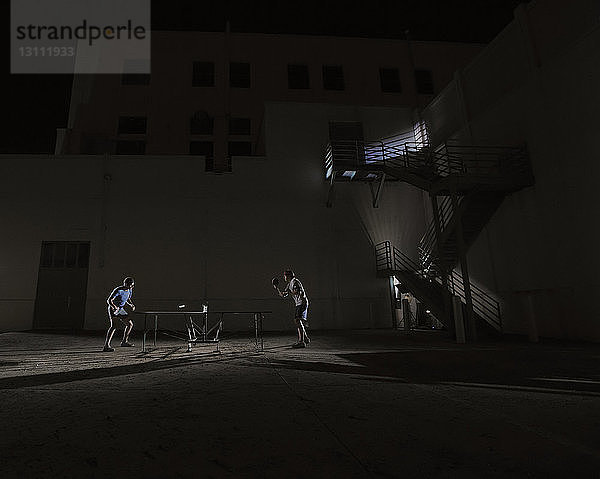 Seitenansicht von Tischtennis spielenden männlichen Freunden gegen beleuchtetes Gebäude