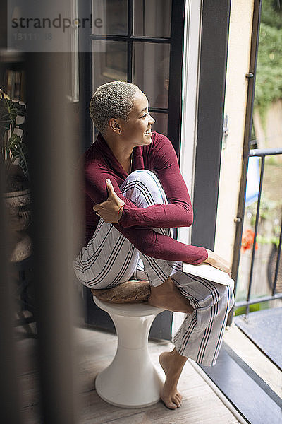 Hochwinkelansicht einer glücklichen Frau  die weg schaut  während sie zu Hause auf einem Hocker sitzt
