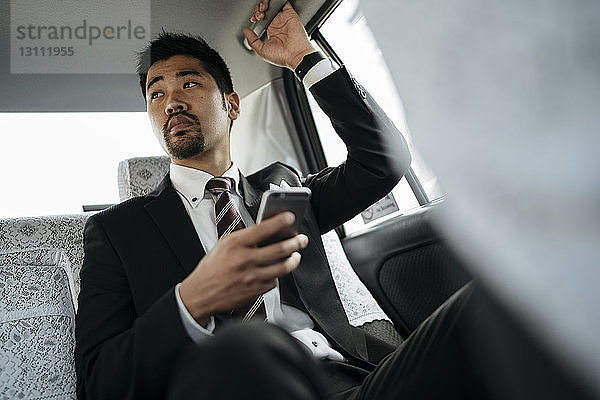 Nachdenklicher Geschäftsmann hält Smartphone in der Hand  während er im Taxi sitzt