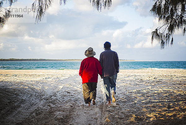 Rückansicht eines älteren Paares  das beim Spaziergang am Sandstrand Händchen hält