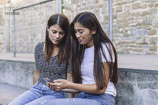Freunde benutzen Mobiltelefon  während sie auf Stufen an der Wand sitzen