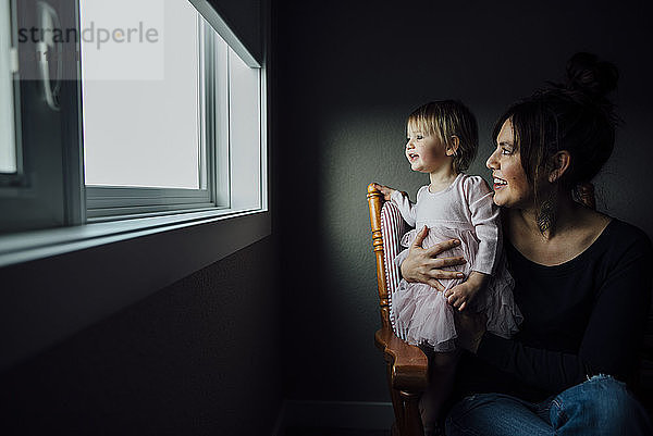 Mutter und Tochter schauen durch das Fenster  während sie zu Hause auf einem Stuhl sitzen