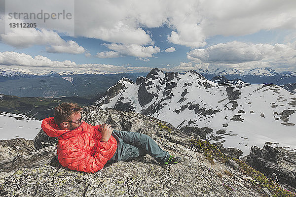 Wanderer in voller Länge mit Smartphone  während er sich im Winter auf dem Berg vor bewölktem Himmel ausruht