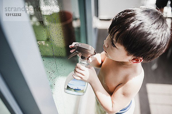 Hochwinkelansicht eines Jungen ohne Hemd  der eine Sprühflasche am Fenster auf dem Balkon hält