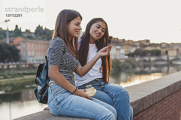 Lächelnde Freundinnen schauen auf die sofortige Druckübertragung  während sie auf einer Stützmauer am Kanal in der Stadt sitzen