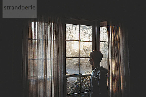 Seitenansicht eines Teenagers  der durch ein Fenster schaut  während er zu Hause steht