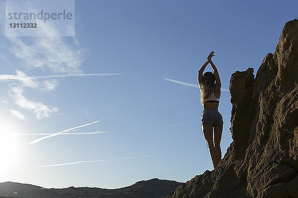 Rückansicht einer Frau mit erhobenen Armen  die auf einer Felsformation vor blauem Himmel steht