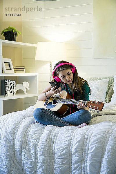 Teenager-Mädchen hört Musik über Smartphone  während sie mit Gitarre im Schlafzimmer sitzt