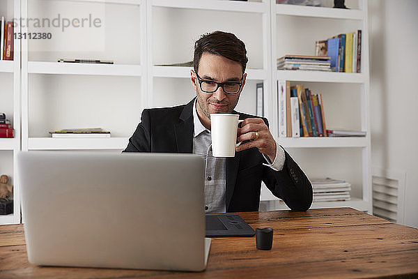 Geschäftsmann hält Kaffeetasse  während er den Laptop am Tisch im Heimbüro benutzt