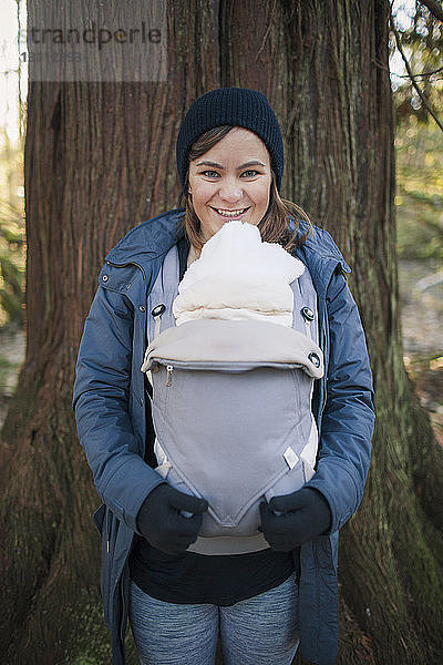 Porträt einer fröhlichen Mutter  die ihren neugeborenen Sohn in einer Babytrage gegen einen Baumstamm im Park trägt