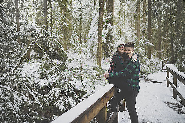 Lachende Frau umarmt Mann  während sie im Winter im Wald im Lynn Canyon Park auf einem Zaun sitzt