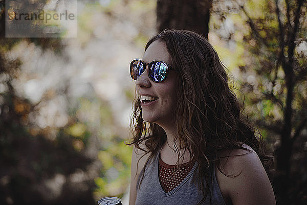 Nahaufnahme einer glücklichen Frau mit Sonnenbrille  die im Wald steht