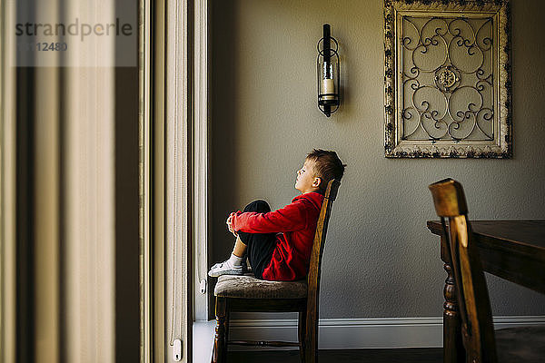 Seitenansicht eines einsamen Jungen  der die Knie umarmt  während er zu Hause auf einem Stuhl sitzt