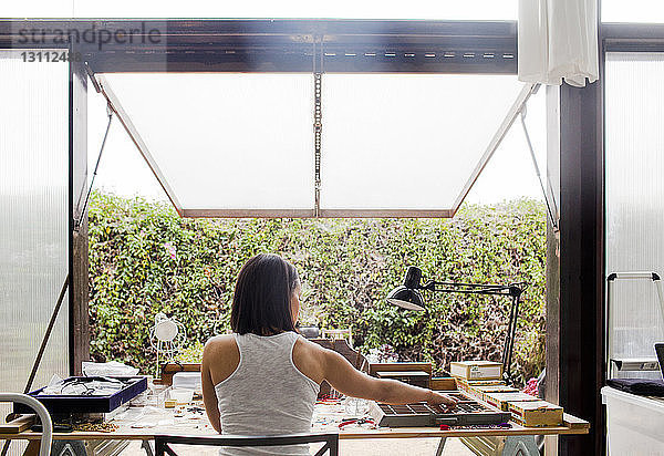 Rückansicht einer am Tisch arbeitenden Frau am Fenster in der Werkstatt