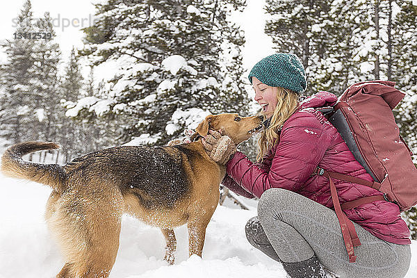 Seitenansicht einer glücklichen Frau mit Rucksack streichelndem Hund  während sie auf einem verschneiten Feld kauert