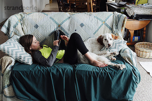 Mädchen benutzt Tablet-Computer  während sie mit Hund zu Hause auf dem Sofa liegt