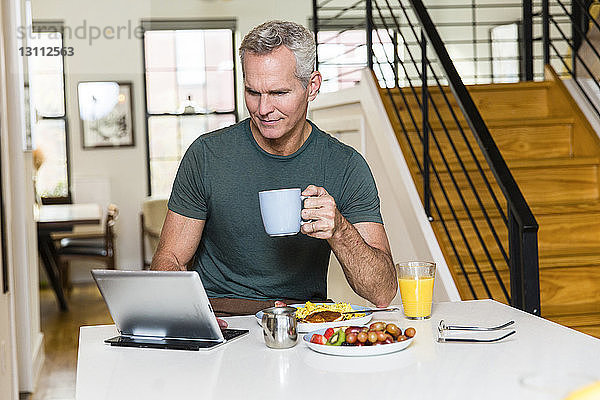 Reifer Mann hält Kaffeetasse  während er einen Tablet-Computer auf dem Esstisch zu Hause benutzt