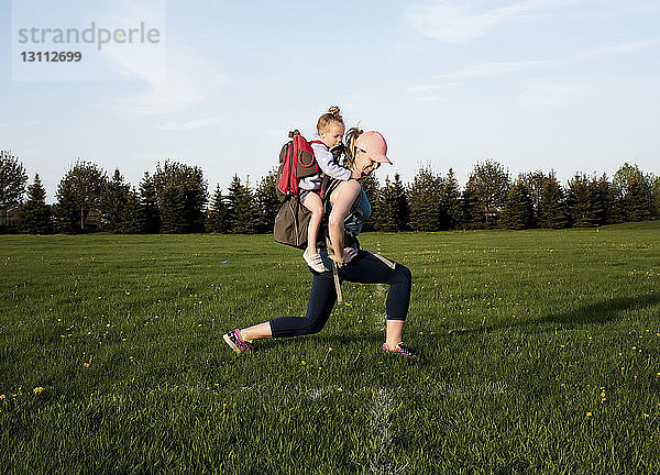 Seitenansicht einer Mutter  die eine Tochter trägt  während sie auf einem Grasfeld gegen den Himmel im Park trainiert
