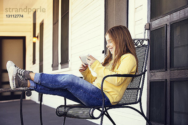 Glückliches Mädchen benutzt Smartphone  während sie auf einem Stuhl sitzt