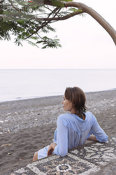 Rückansicht einer nachdenklichen Frau  die sich entspannt  während sie am Strand gegen den Himmel sitzt