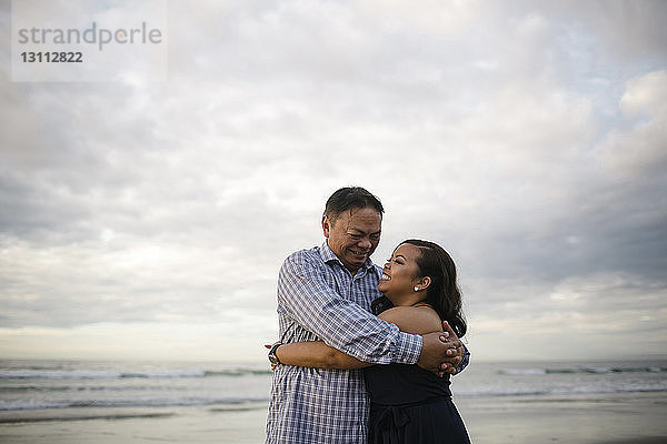 Glückliche Frau umarmt Vater  während sie am Strand vor bewölktem Himmel steht