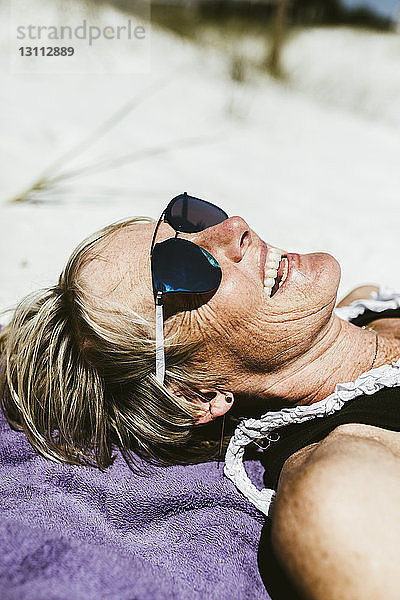 Fröhliche  reife Frau mit Sonnenbrille  die bei Sonnenschein am Strand liegt