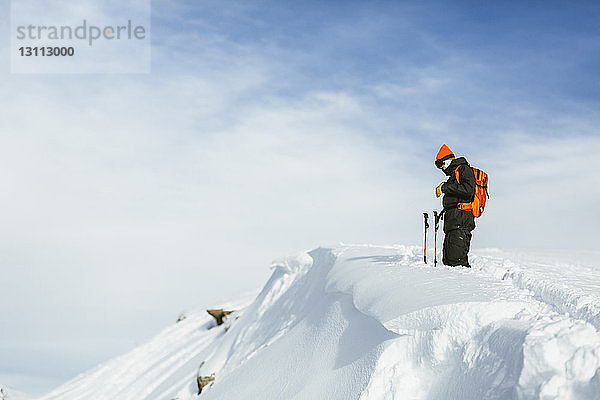 Seitenansicht eines Wanderers mit Rucksack und Skistöcken  der auf einem schneebedeckten Berg vor bewölktem Himmel steht