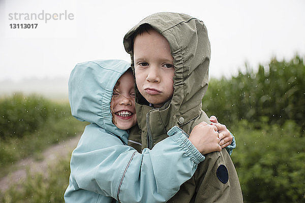 Porträt eines traurigen Bruders  der von einer glücklichen Schwester umarmt wird  während er in der Regenzeit vor Pflanzen steht